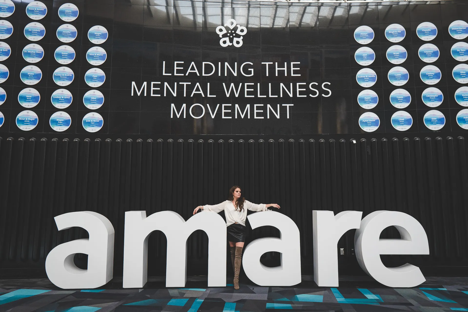 Lindsey Elmore leaning against giant white amare letters on the convention floor, beneath a wall reading Leading the Mental Wellness Movement