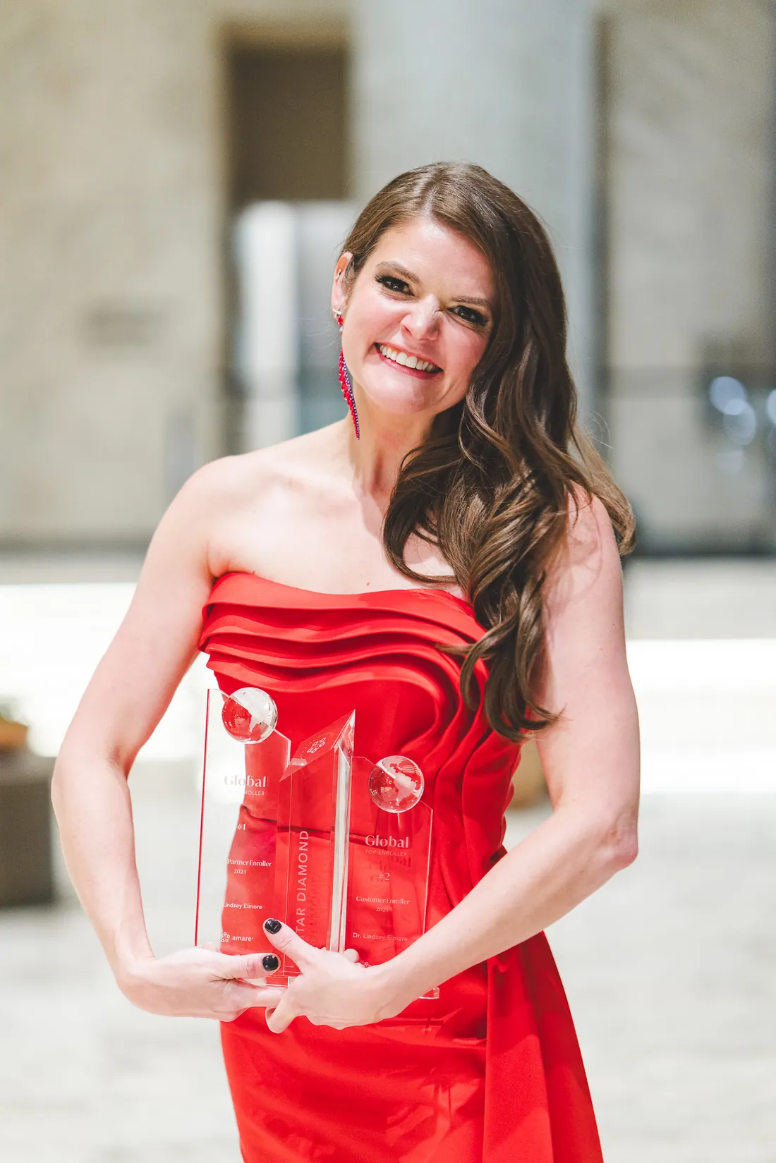 Lindsey Elmore smiling in a red gown, holding two crystal awards