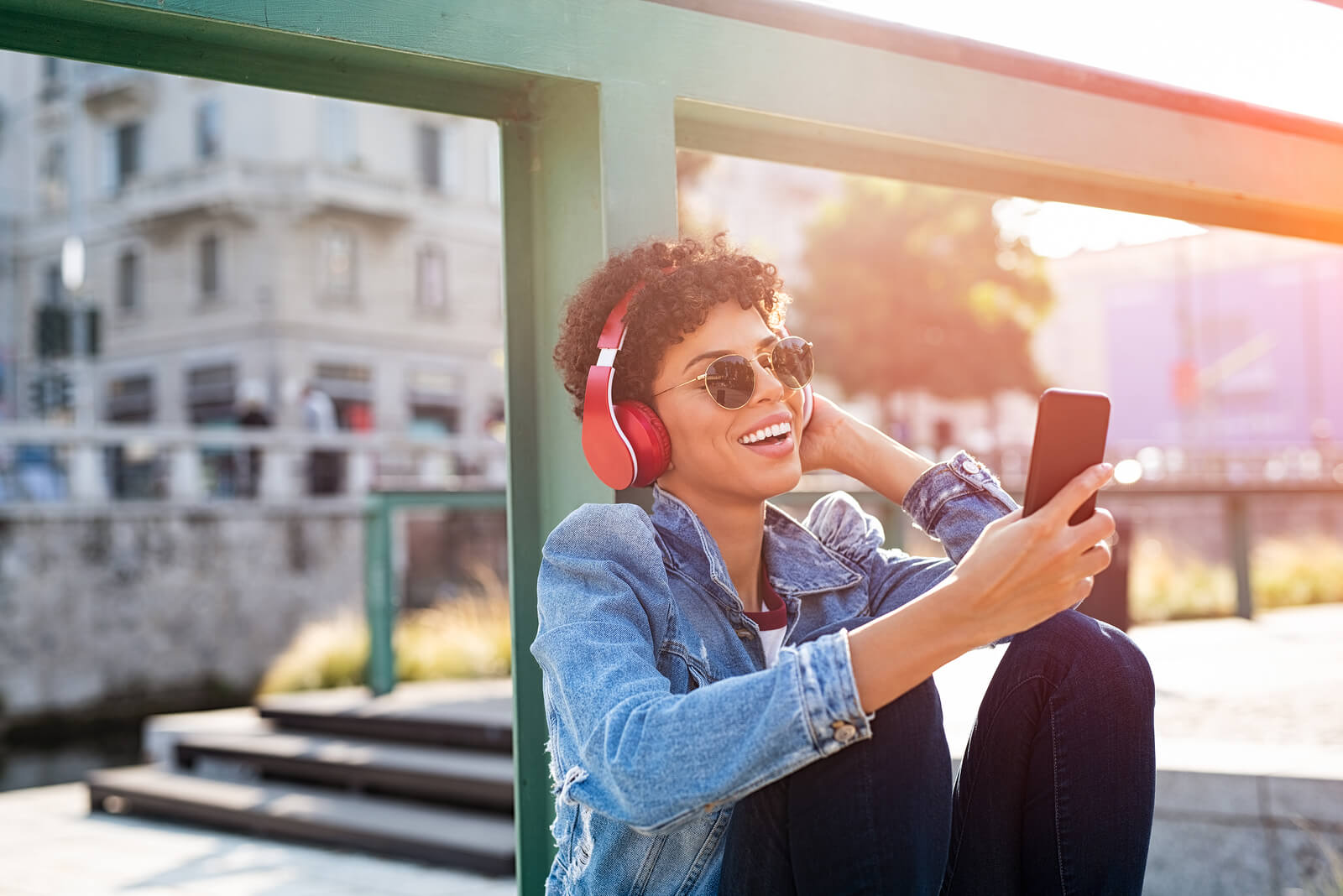 Young beautiful woman holding smartphone and listening to music with wireless headphones. Cheerful smiling girl with red headphones changing music track on phone. Carefree urban teen enjoying songs.